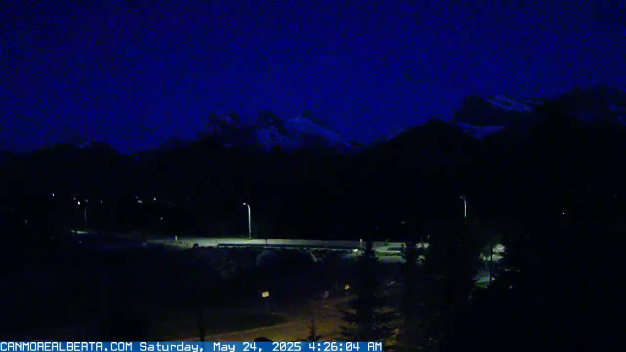 A nighttime view of dark mountains under a dark blue sky, with a few dimly lit streetlights visible in the foreground.