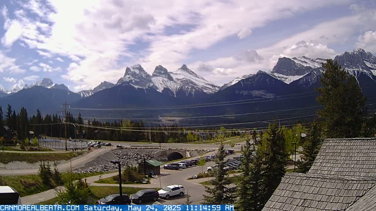 A partly cloudy day reveals a mountain range with snow-capped peaks, overlooking a parking lot and roadway nestled among evergreens.