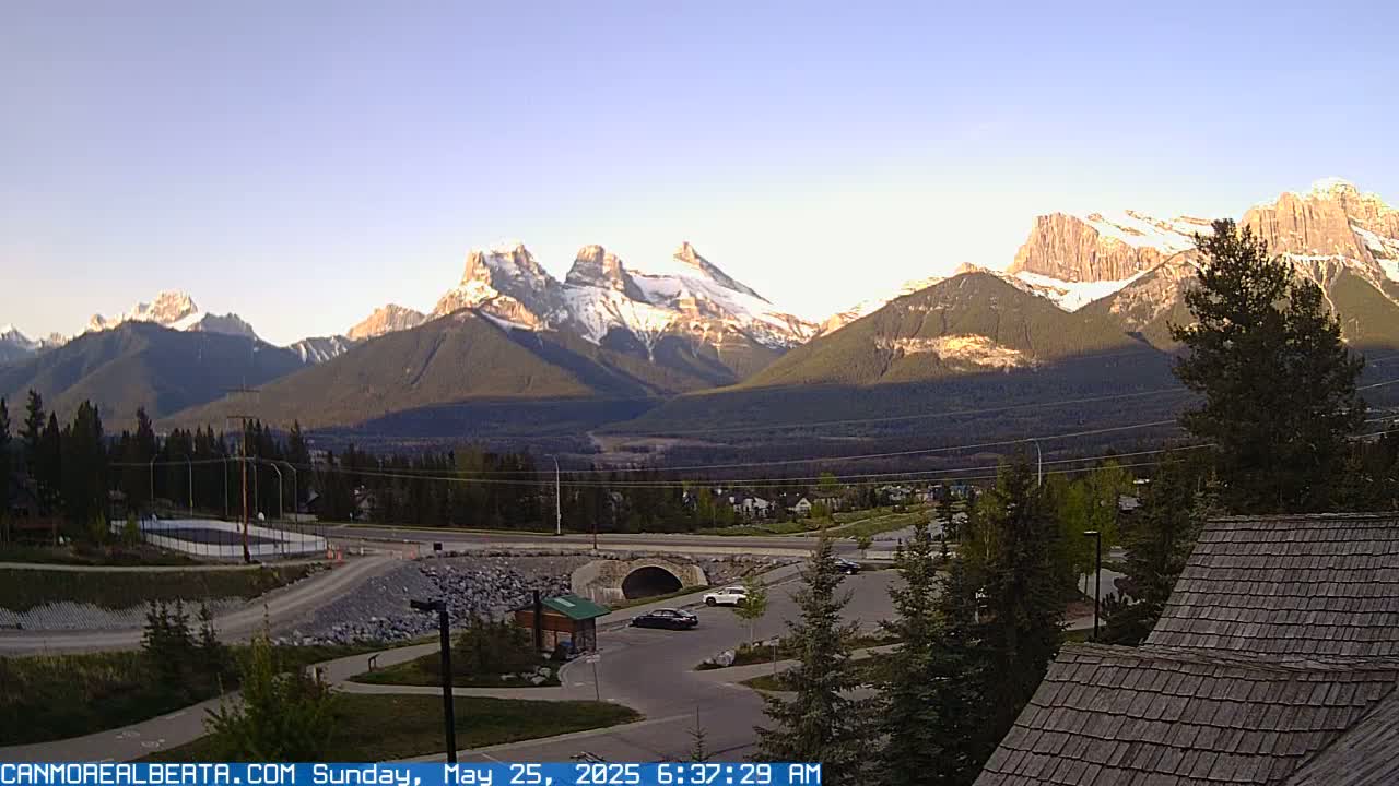 A clear sunrise illuminates snow-capped mountains behind a valley with roads, trees, and a parking lot.