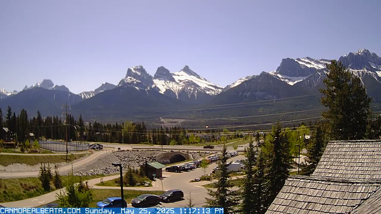 A sunny, outdoor scene shows snow-capped mountains in the background, with a parking lot, road, and some buildings in the foreground.