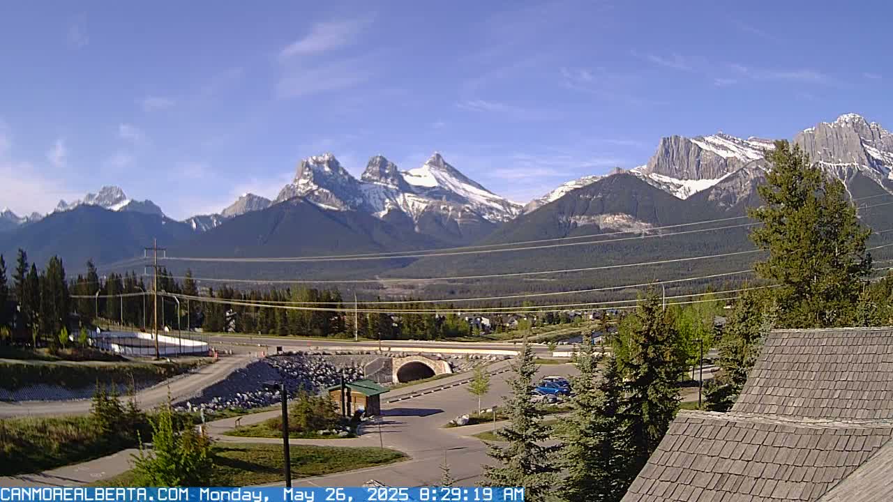 A sunny day reveals a mountain range with snow-capped peaks, overlooking a valley with a road, bridge, and some buildings.