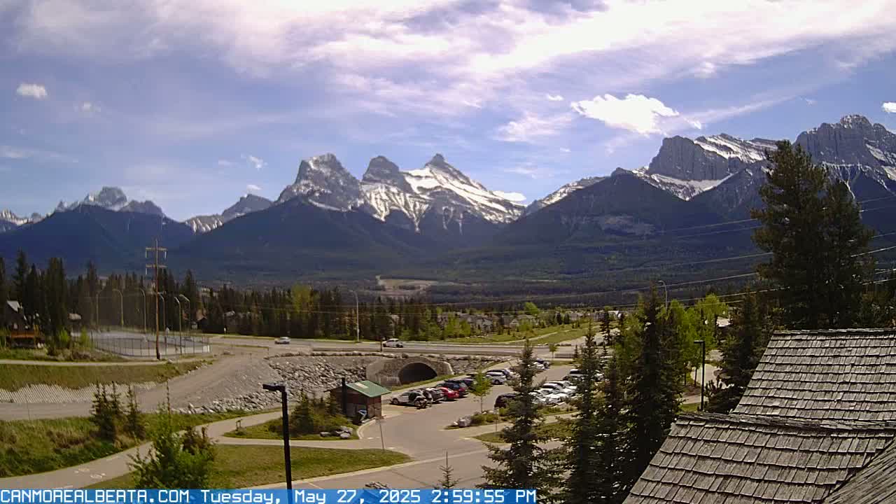 A partly cloudy day reveals a mountain range with snow-capped peaks in the background, overlooking a valley with a parking lot, a road, and some buildings in the foreground.