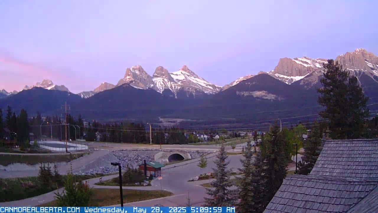 A view of snow-capped mountains at dawn, overlooking a town with a road, park, and buildings.