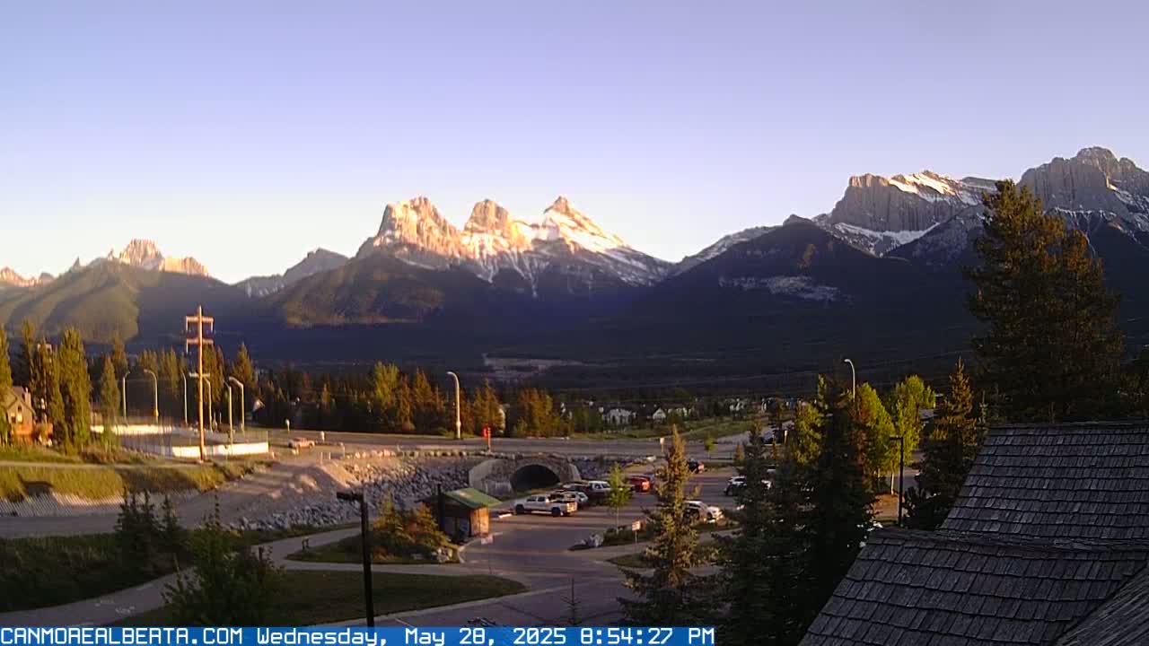 A mountain range with snow-capped peaks is visible in the distance under a clear evening sky, with a town and parking lot in the foreground.