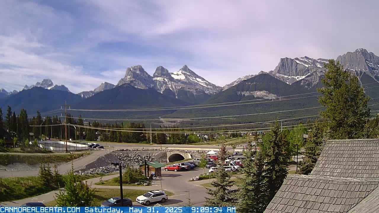 A partly cloudy day reveals a mountain range with snow-capped peaks in the distance, overlooking a parking lot and roadway with vehicles, and some trees and buildings in the foreground.