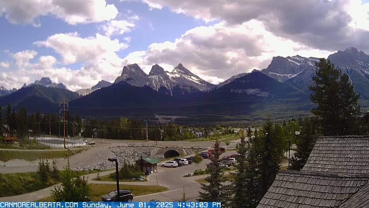 A partly cloudy day reveals a mountain range with snow-capped peaks, a valley with a small town, a parking lot with several cars, and a portion of a building with a shingled roof.