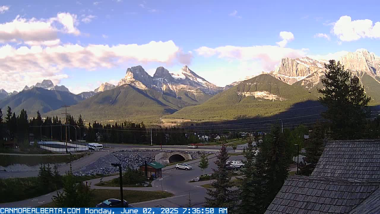A partly cloudy morning reveals a mountain range with snow-capped peaks, seen from a parking lot with several vehicles.