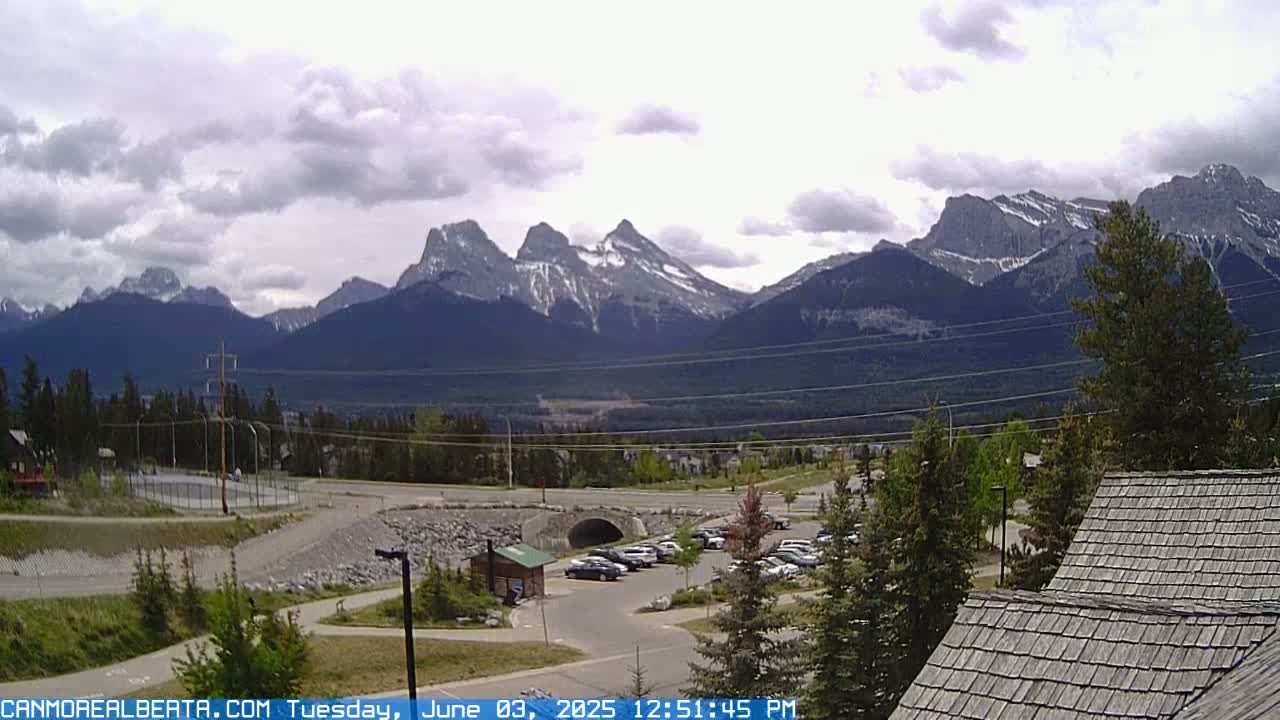 A parking lot and surrounding landscape are visible in the foreground, with a range of snow-capped mountains under a partly cloudy sky in the background.
