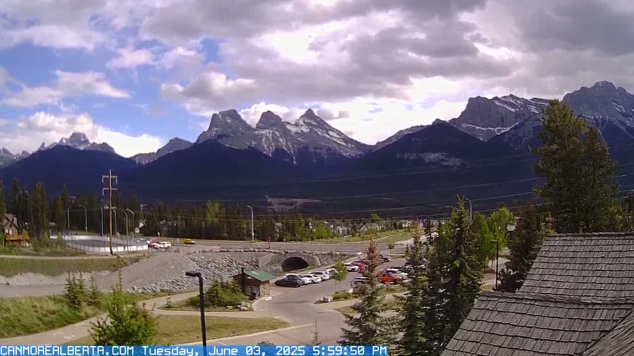 A partly cloudy day reveals a mountain range with snow-capped peaks in the background, overlooking a town with a parking lot, road, and bridge in the foreground.