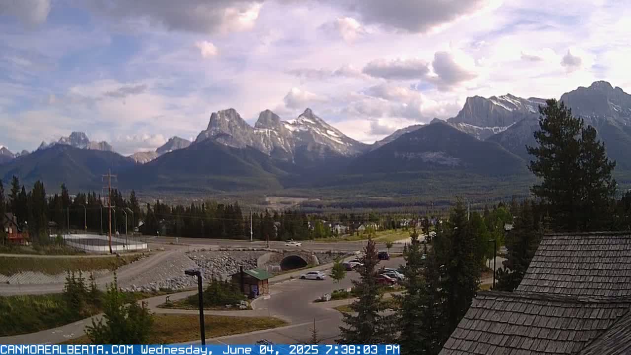 A partly cloudy day reveals a mountain range with some snow cover, visible from a parking lot and surrounding area with trees and a small building.