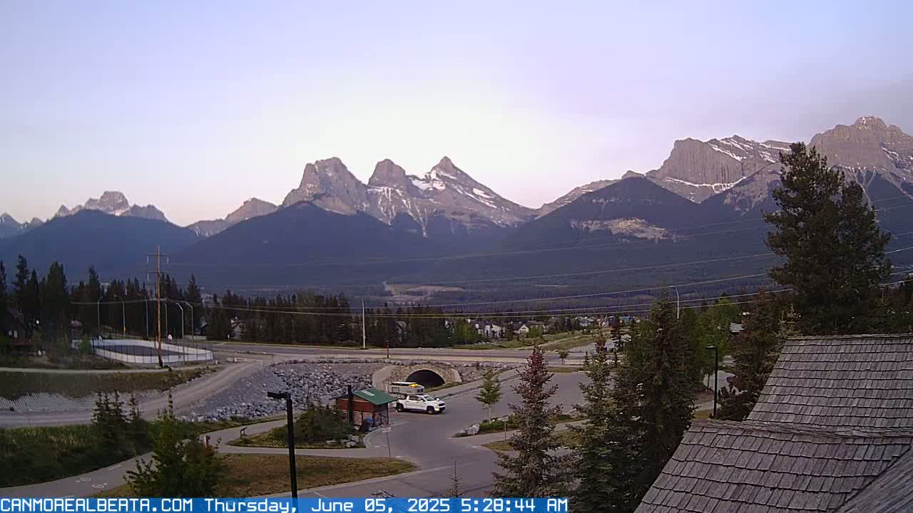 A mountain range under a clear, dawn sky is visible from a parking lot with a single vehicle, nestled amongst trees and buildings.