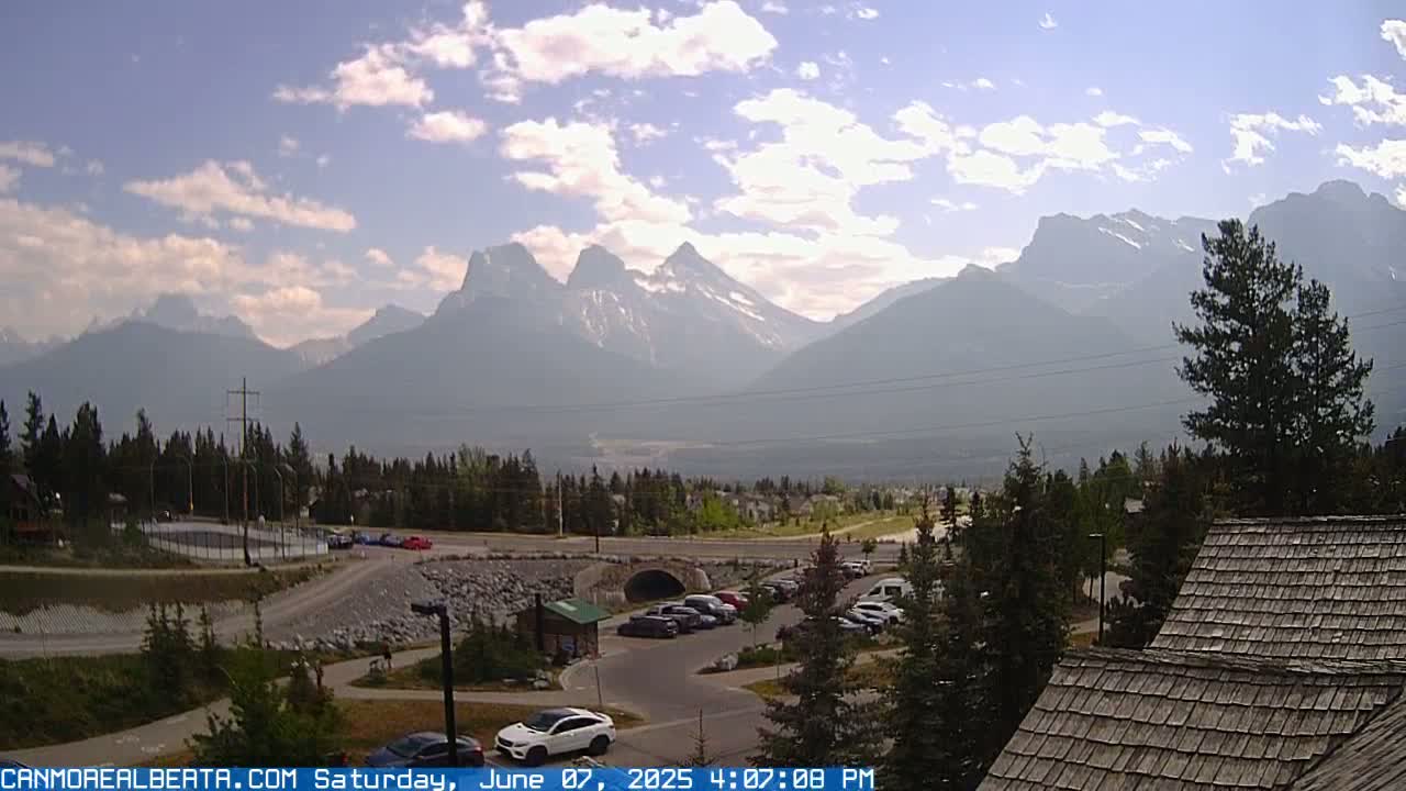 A partly cloudy day reveals a mountain range in the distance, with a parking lot, road, and buildings in the foreground.