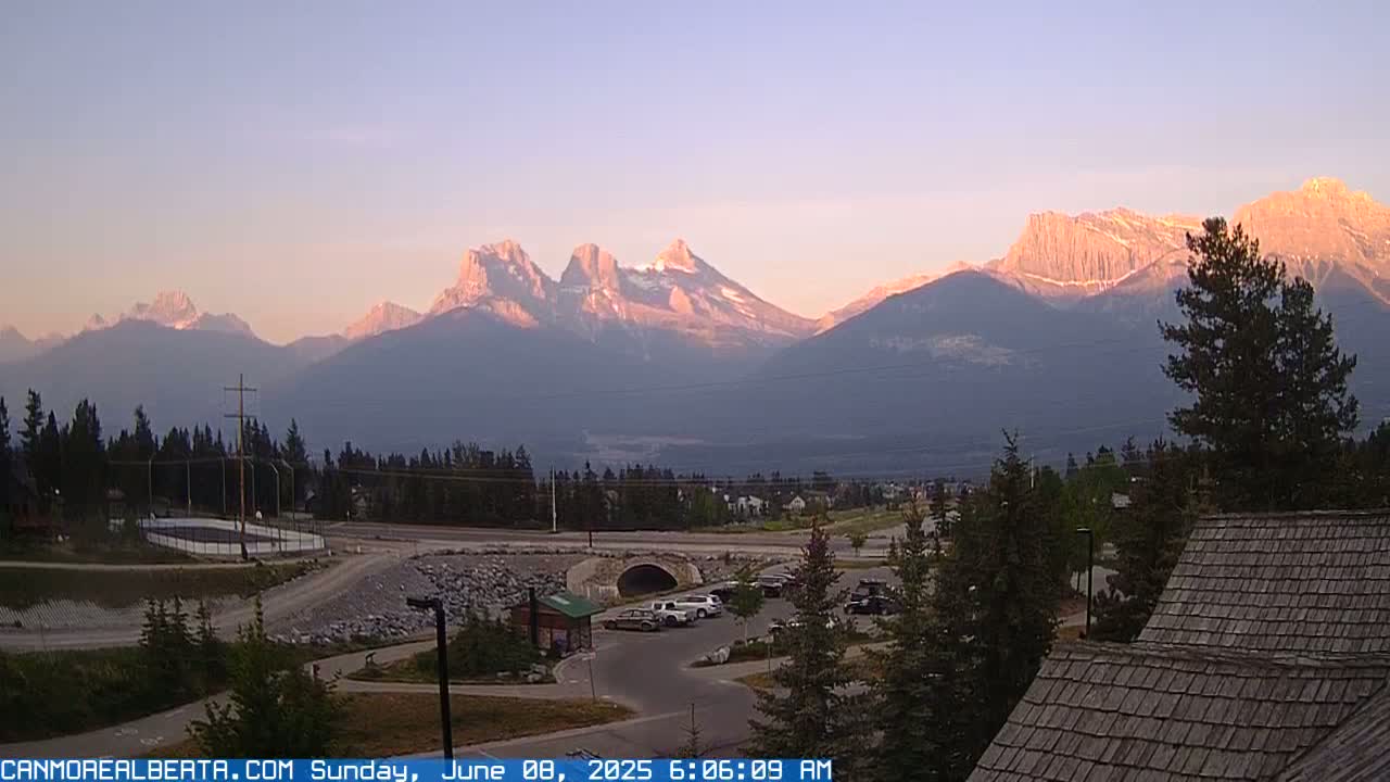 A sunrise illuminates a mountain range behind a small town with a parking lot and a building with a wood-shingle roof.