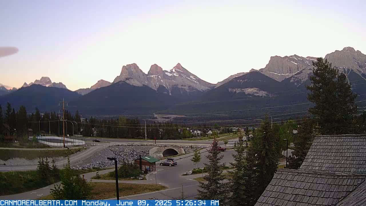A mountain range at sunrise is seen from a high vantage point overlooking a valley with a road, some buildings, and a parking lot.