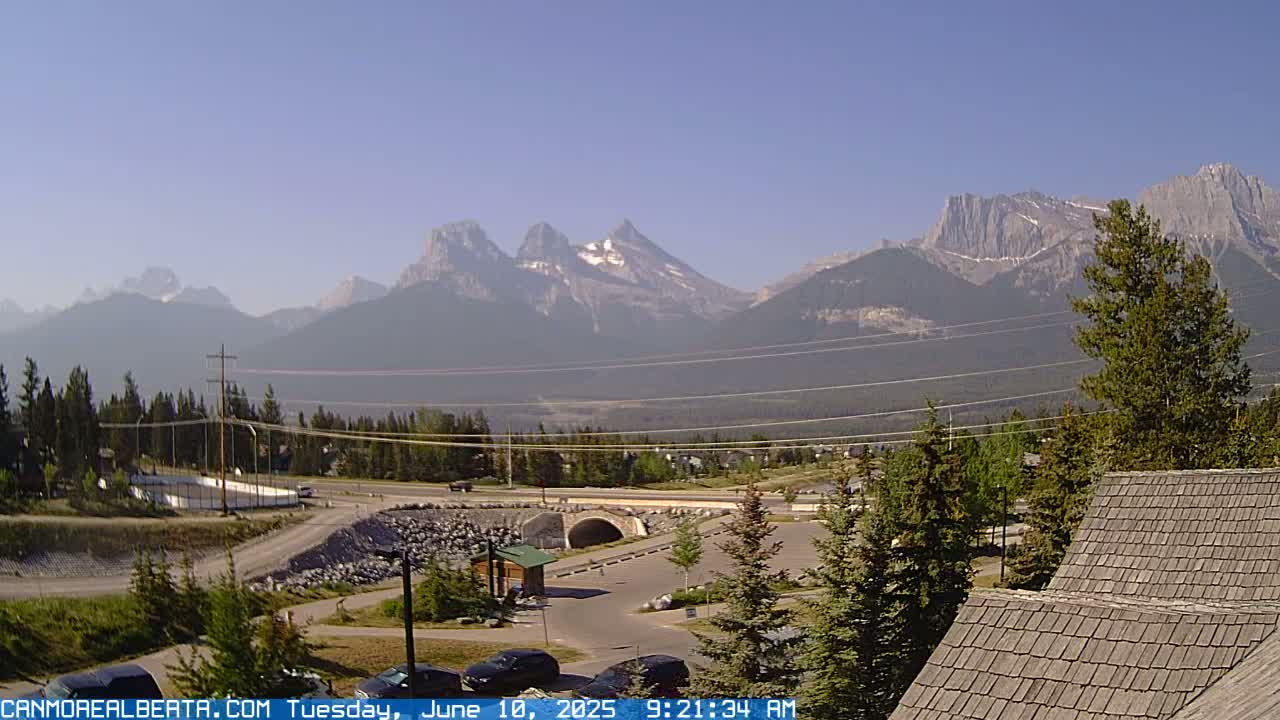 A clear, sunny day reveals a mountain range in the distance, with a town, road, and bridge in the foreground, partially obscured by evergreen trees and a portion of a building's roof.
