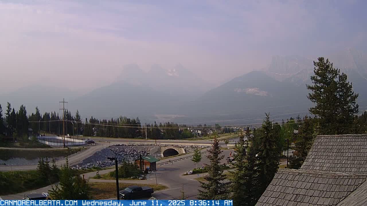 A hazy, smoky view of a mountain range in the distance, overlooking a town with a road, parking lot, and some buildings, including a section of a roof in the foreground.