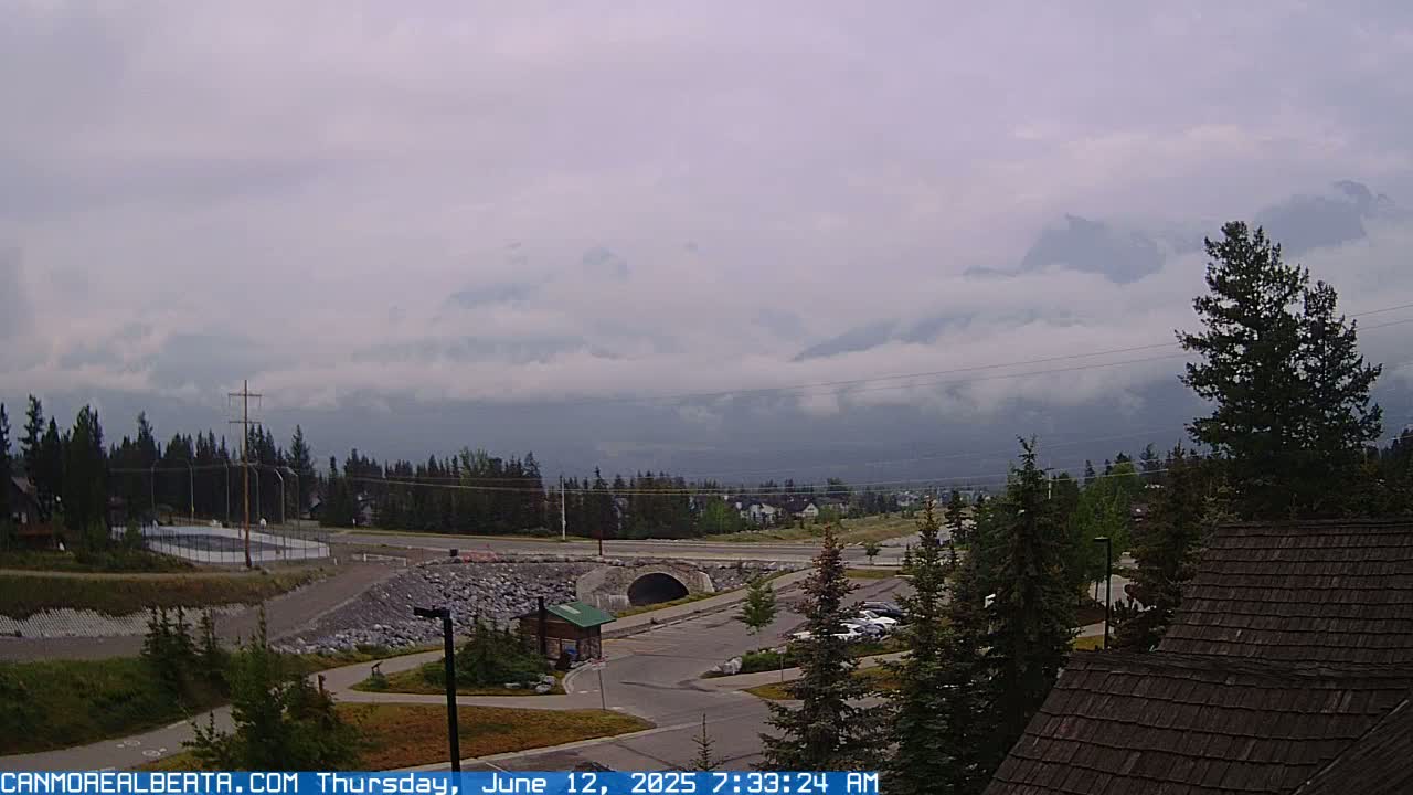 A cloudy, overcast day reveals a mountain range partially obscured by clouds, a small town nestled in a valley with a roadway and parking lot, and a portion of a building with a shingled roof in the foreground.