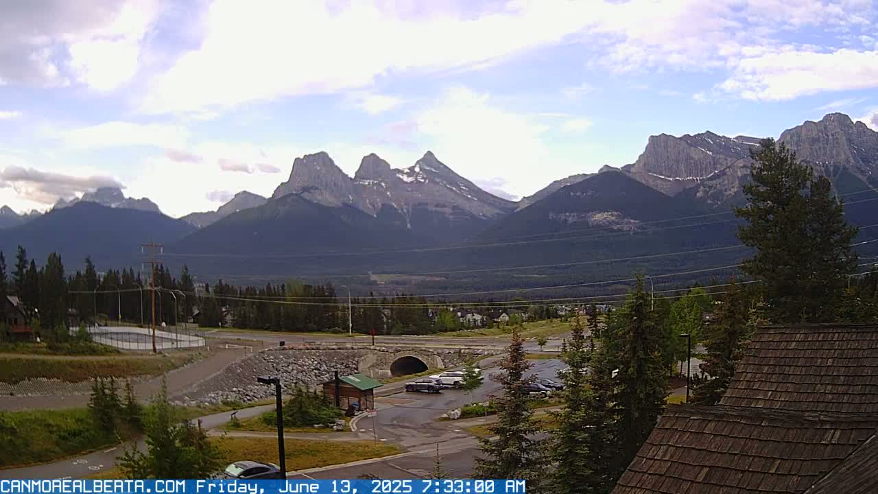 A partly cloudy sky overlooks a mountain range, a parking lot with several cars, a small tunnel, and some buildings.