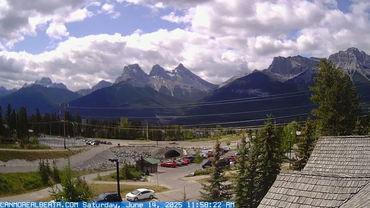 A partly cloudy day reveals a mountain range in the background, with a parking lot, roadway, and some buildings in the foreground.