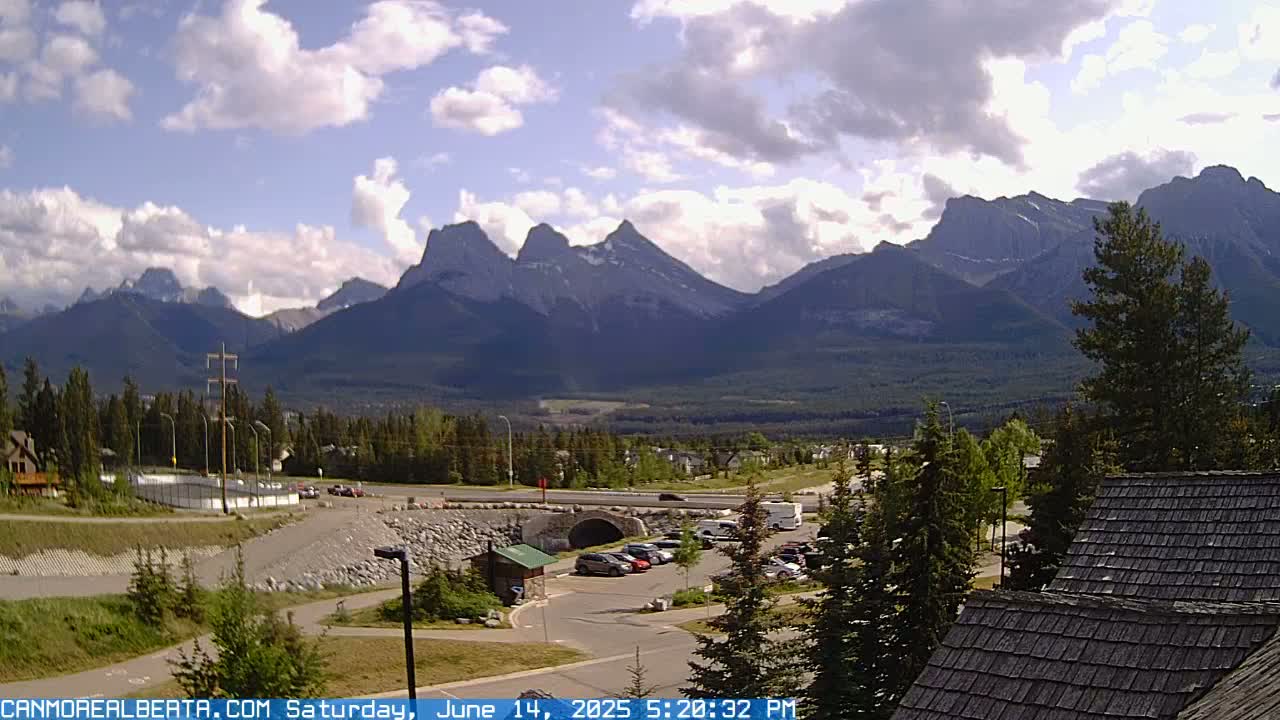 A partly cloudy day reveals a mountain range in the background, with a parking lot, roadway, and some buildings in the foreground.