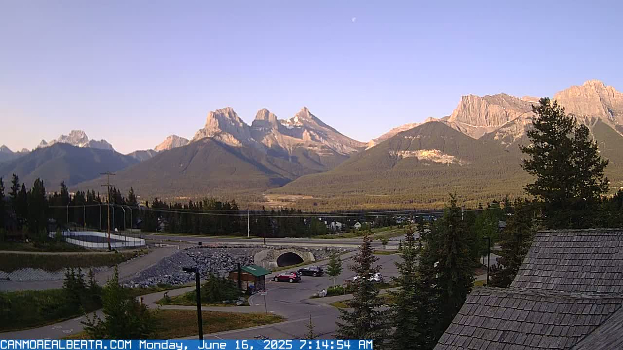 A clear, sunny morning view shows a valley with a road, parking lot, and several buildings nestled in front of a large mountain range.