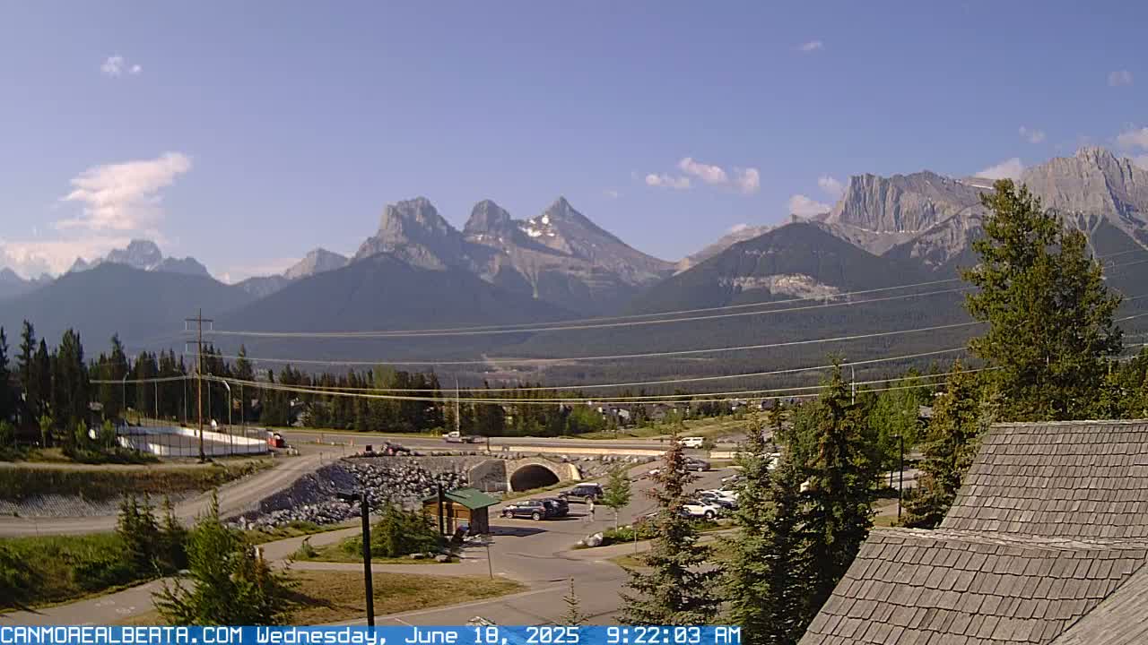 A partly cloudy day reveals a mountain range in the distance, with a parking lot, road, and small buildings in the foreground.