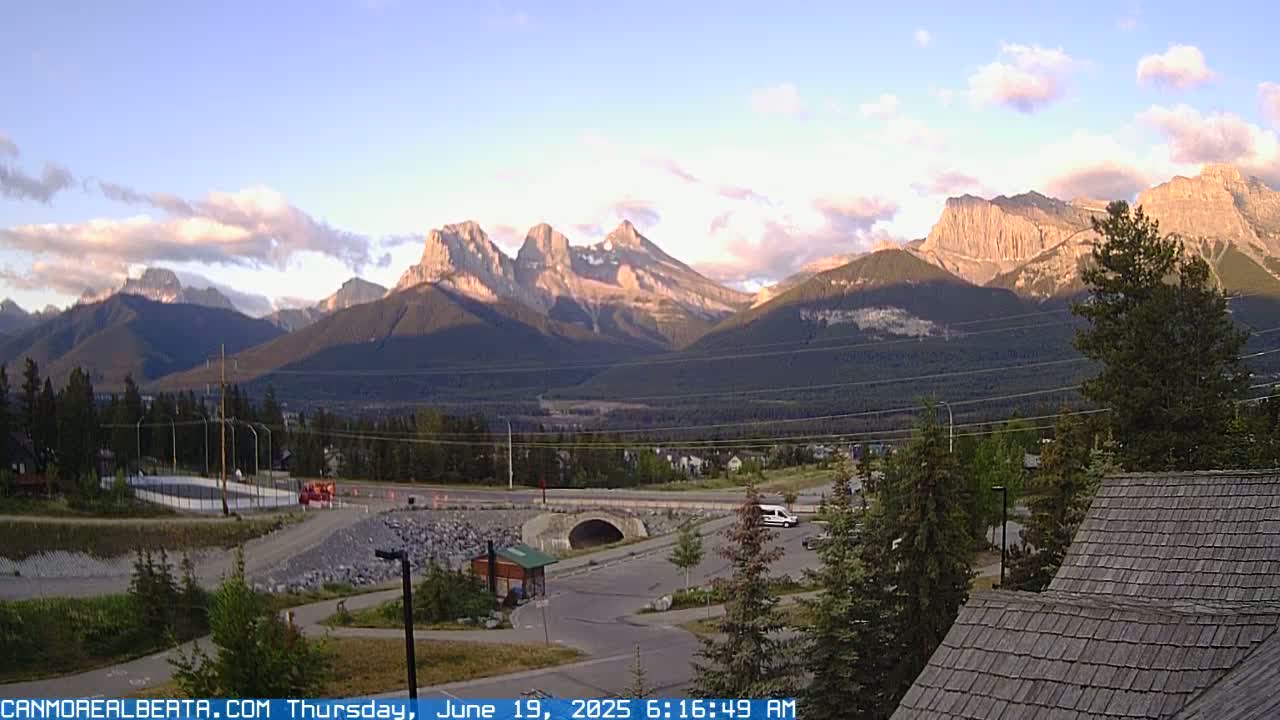 A mountain range is visible under a partly cloudy sky at sunrise, overlooking a valley with a road, a parking lot, and some buildings.