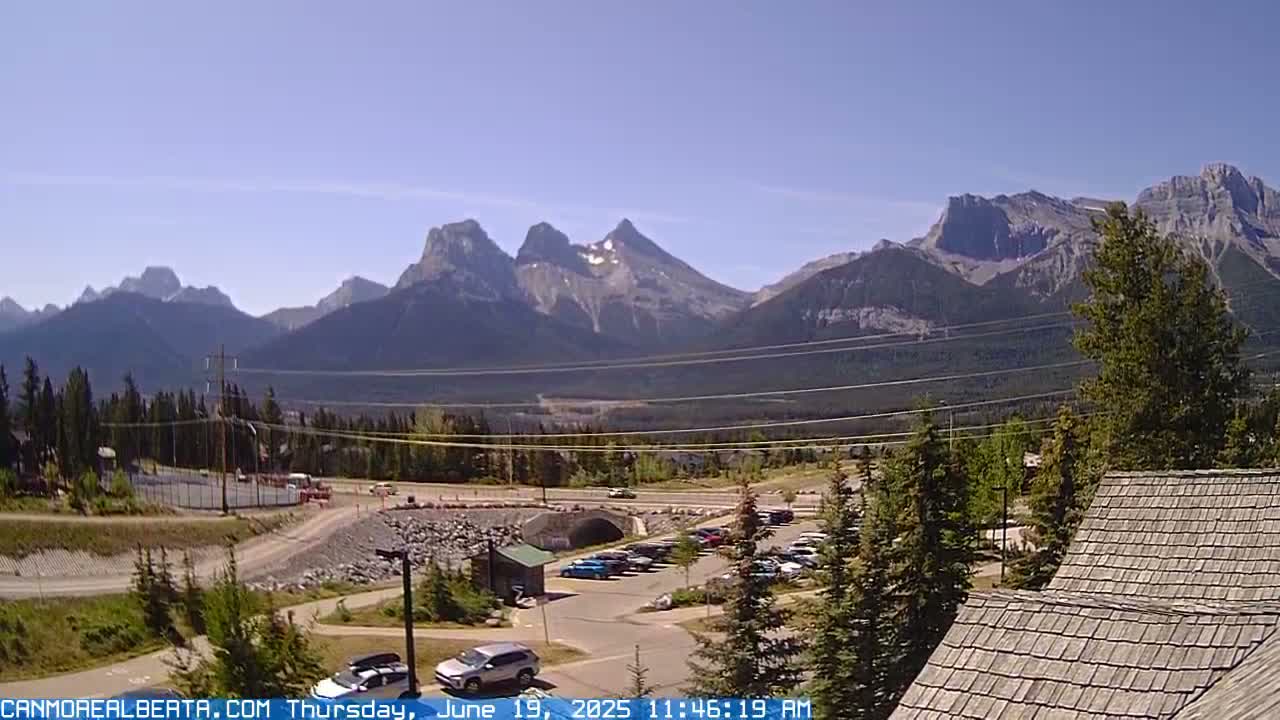 A parking lot and roadway are nestled among evergreen trees, with a mountain range in the background under a clear, sunny sky.