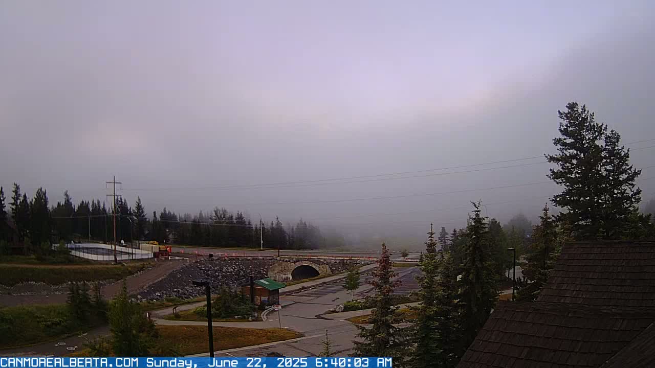 A foggy morning scene shows a road, a stone bridge, a parking lot, and evergreen trees, with a portion of a building's roof in the foreground.