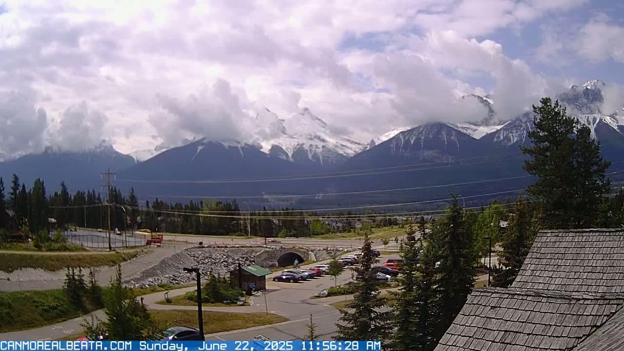 A partly cloudy day reveals snow-capped mountains in the distance, overlooking a parking lot, a roadway, and evergreen trees in the foreground.