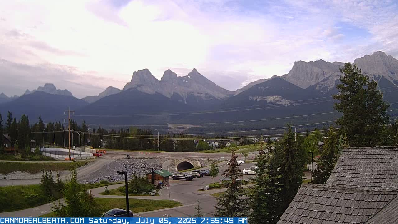 A partly cloudy morning view shows a mountain range in the background, a parking lot with several cars, a roadway with construction, and a portion of a building with a shingled roof in the foreground.