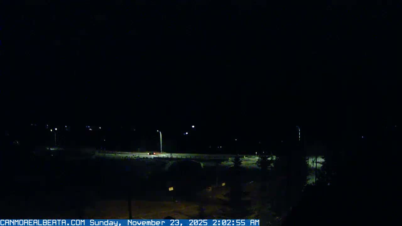 A dark, clear night scene shows a road or bridge illuminated by streetlights, with a vehicle's headlights visible on the road and distant lights dotting the landscape.