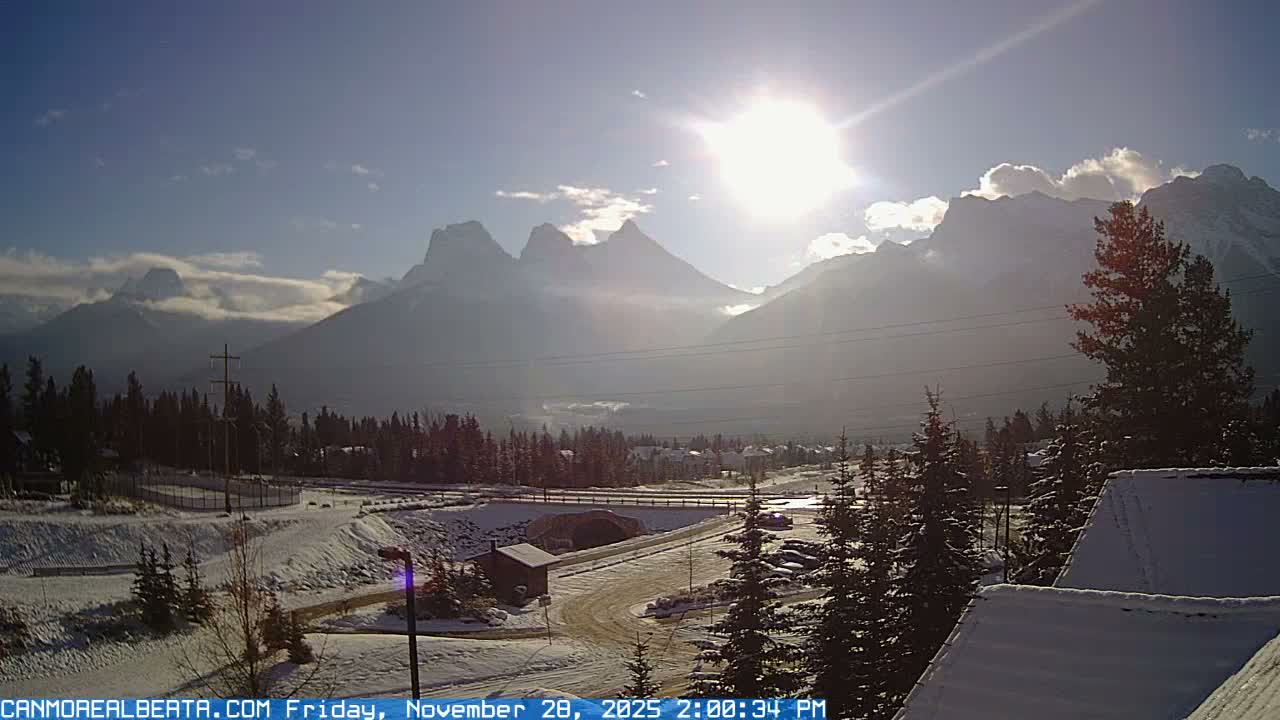 A sunny winter day reveals a snow-covered mountain valley with a winding road, coniferous trees, and distant peaks partially veiled by a bright sun and light haze.
