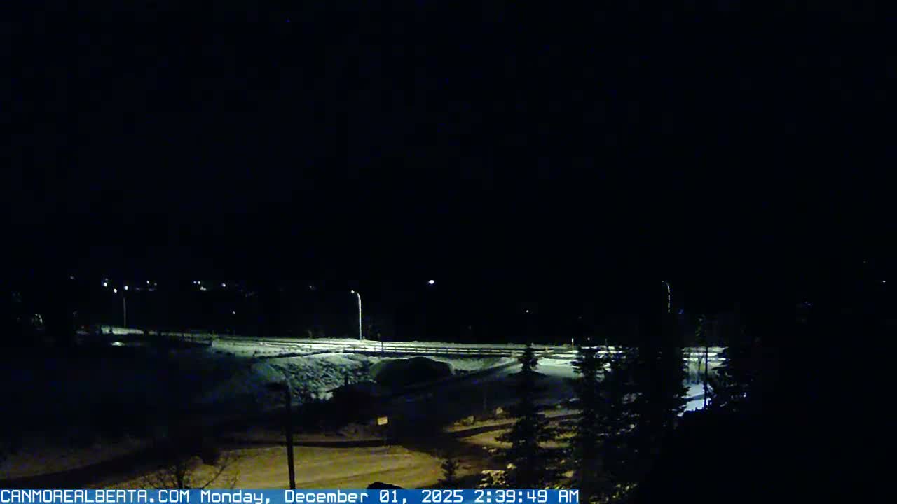 A clear, cold winter night scene reveals a snow-covered landscape with a highway illuminated by streetlights and distant town lights under a dark sky.