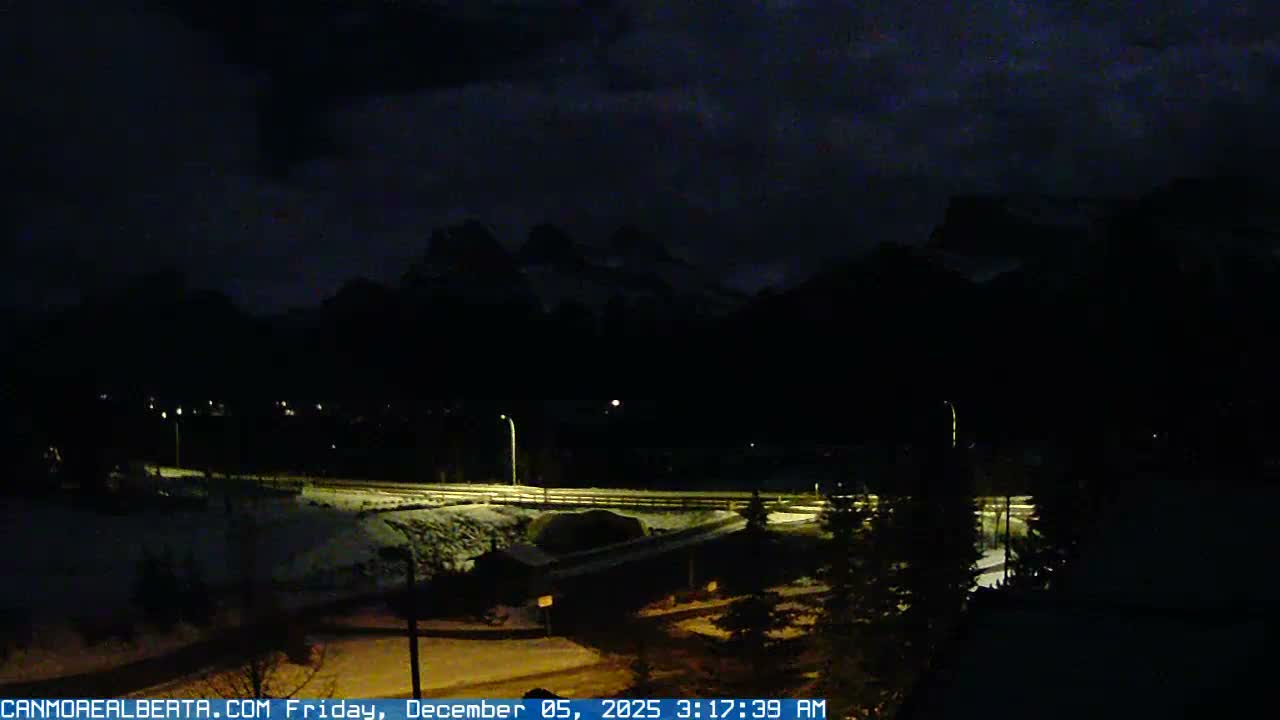 A snowy, mountainous landscape at night is illuminated by streetlights along a road under a dark, cloudy sky.