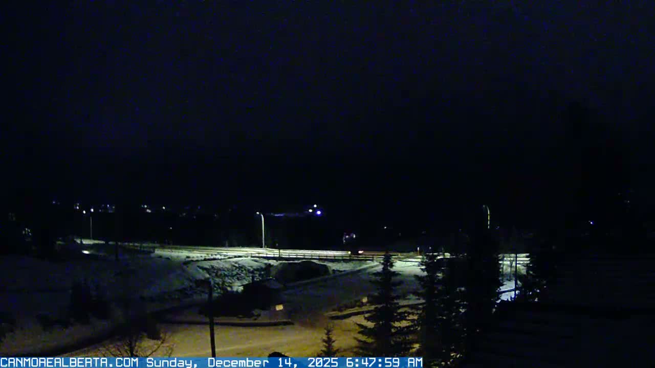 A snowy, mountainous landscape at night is illuminated by streetlights along a road under a dark, cloudy sky.
