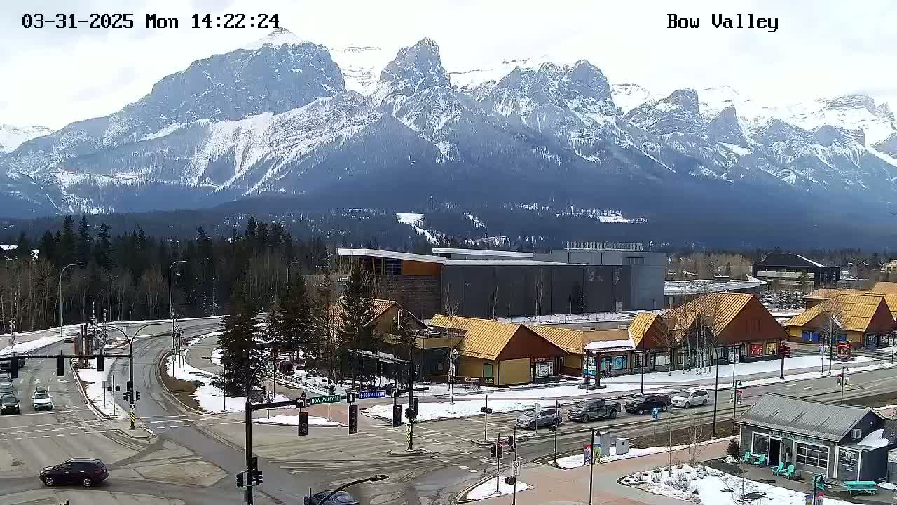 A snow-dusted mountain range overlooks a valley town with roads, buildings, and sparse vehicle traffic under a cloudy sky.