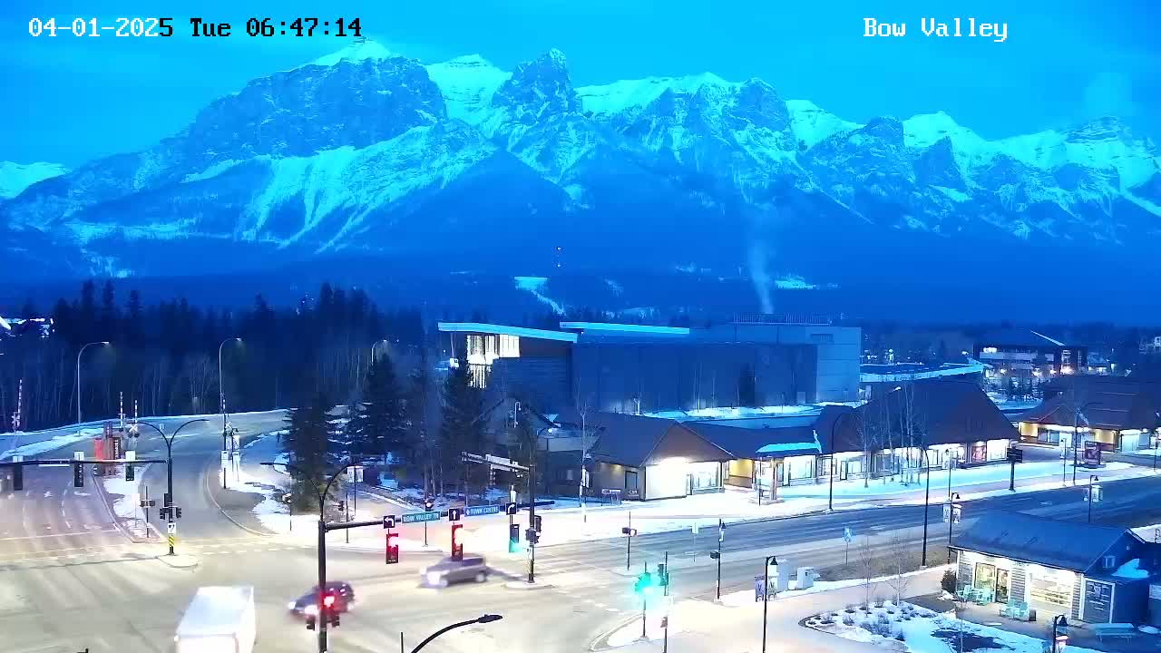 A snow-covered town with a four-way intersection and buildings sits at the base of a large, snow-capped mountain range under a twilight blue sky.