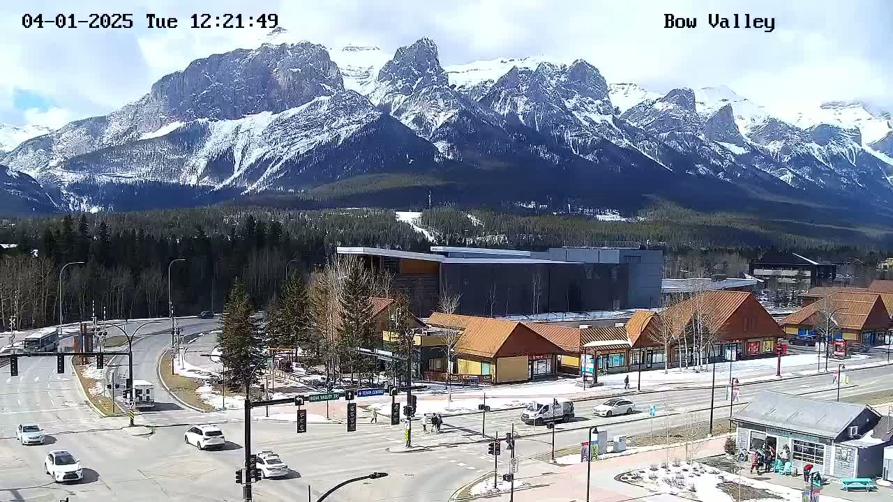 A partly sunny day shows a mountain range with snow-capped peaks behind a town with buildings and vehicles, at a road intersection.