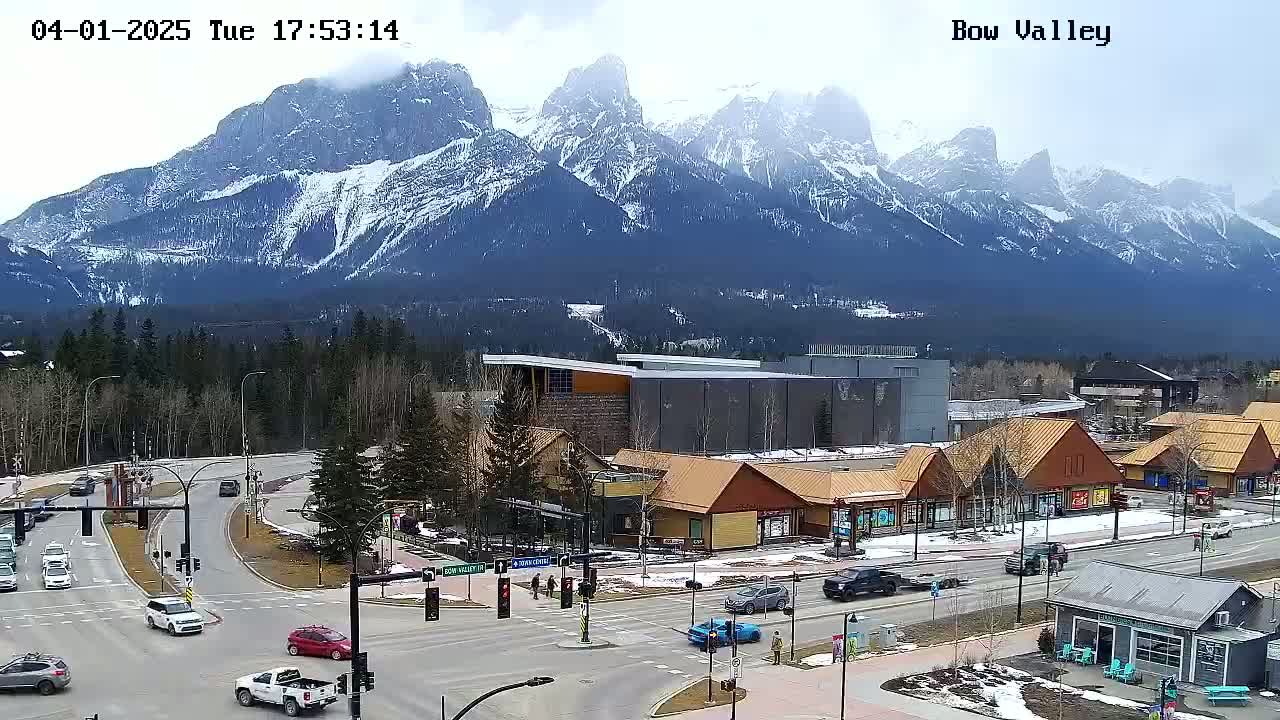 A partly cloudy day reveals snow-capped mountains in the background, overlooking a town with roads, buildings, and cars.