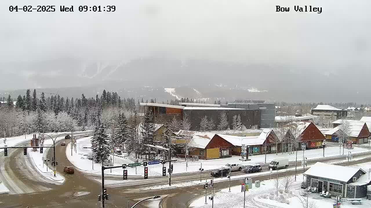 A snow-covered town scene shows a road intersection with several cars, surrounded by snow-covered buildings and trees, under a gray, overcast sky.