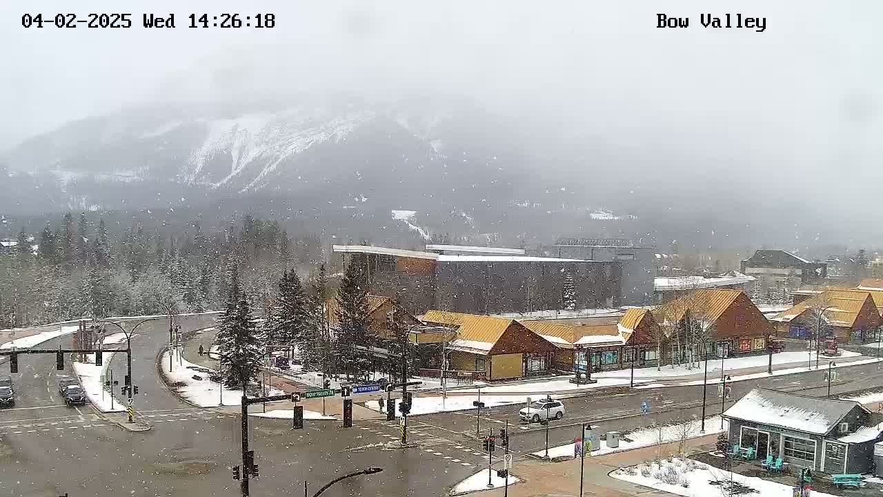 A snow-covered mountain range is visible in the background of a lightly snowing town scene showing a street intersection with several buildings, including some shops with pitched roofs.