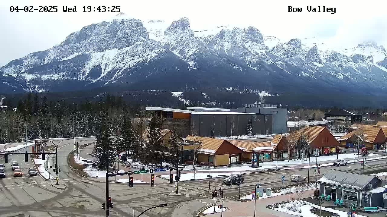 A snow-covered mountain range is visible in the background of a town scene with buildings, roads, and cars, on a cloudy day.