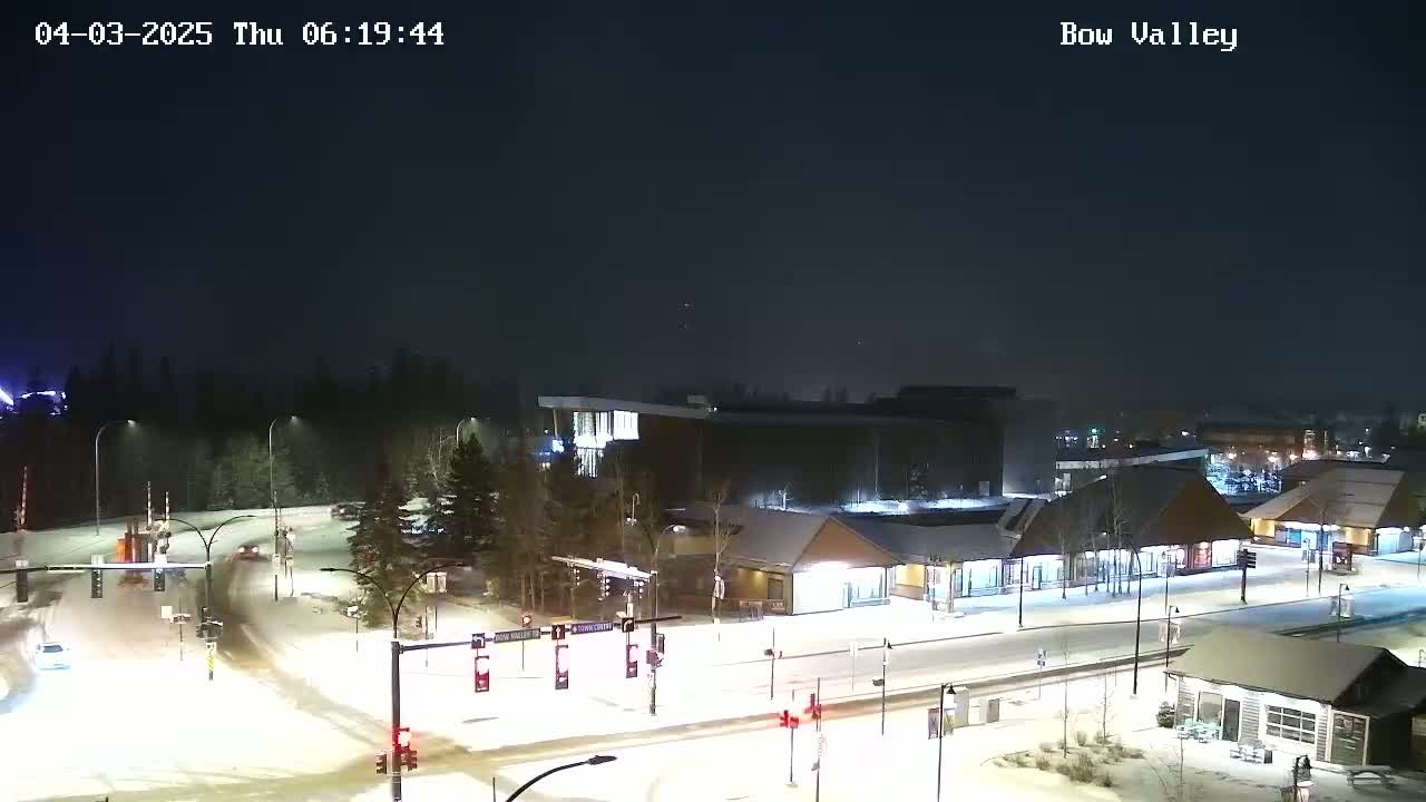 A snow-covered town square at night, illuminated by streetlights, with several buildings and vehicles visible.