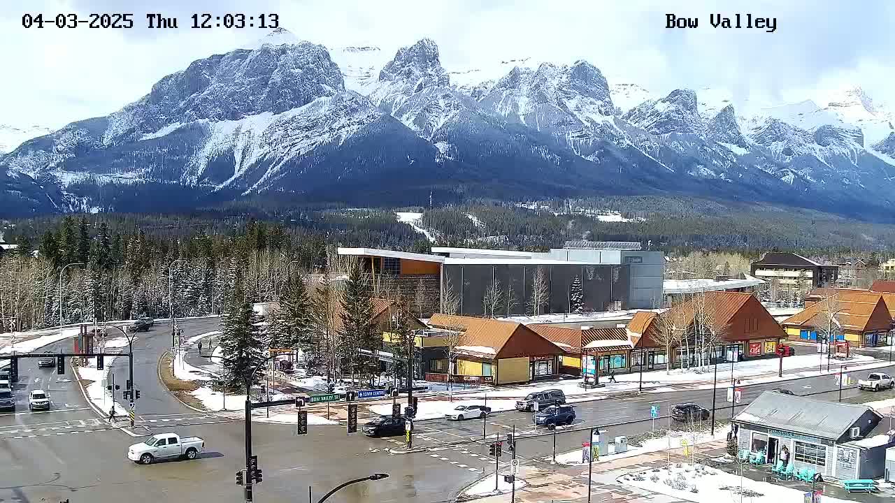 A mountain range capped with snow overlooks a town with buildings and roads partially covered in snow under partly cloudy skies.