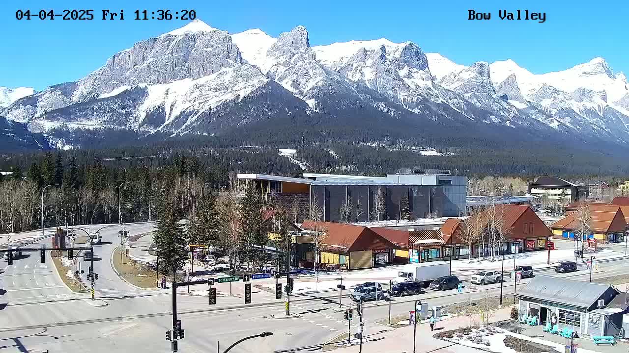 A sunny day reveals a mountain range capped with snow behind a town with buildings and roads, featuring a few cars and pedestrians.
