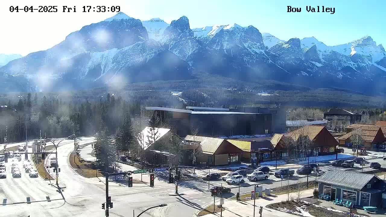 A sunny day reveals a mountain range in the background, overlooking a town with buildings, roads, and vehicles, some snow is visible on the ground and mountains.