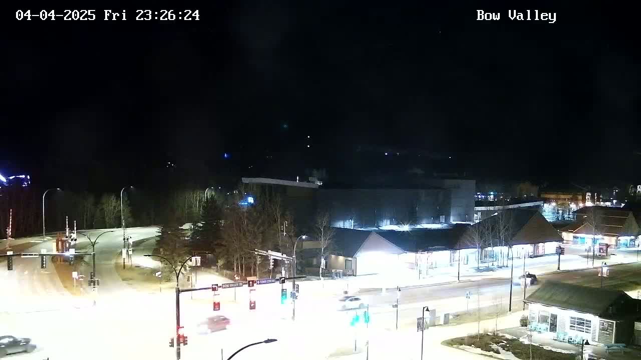 A nighttime aerial view of a snow-covered intersection with several buildings and cars, under a clear, dark sky.