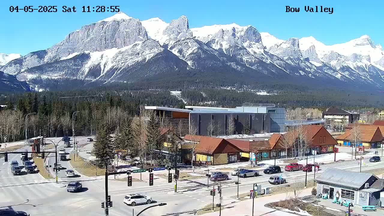 A sunny day reveals a mountain range capped with snow overlooking a town with a crossroads and various buildings, including shops and a larger structure.