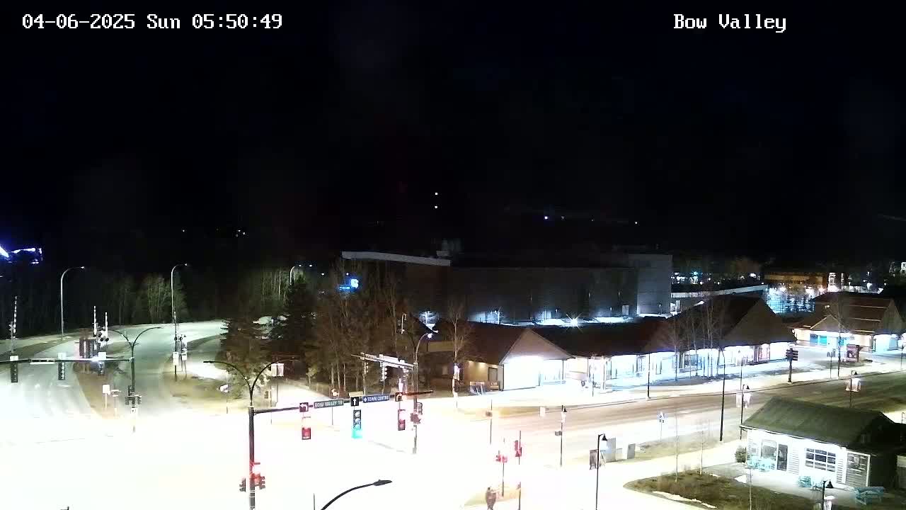 A nighttime, snow-covered intersection with illuminated buildings and streetlights.